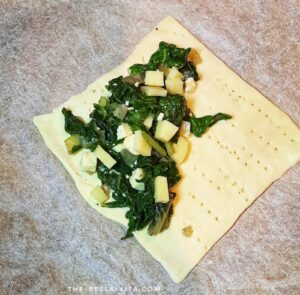 Close-up of spinach and ricotta filling being placed on puff pastry, ready to be folded for a savory Italian pastry. The Bella Vita.