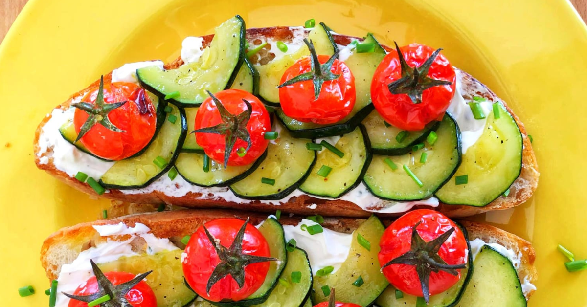 Delicious roasted tomato and zucchini bruschetta on toasted bread with creamy cheese, garnished with fresh chives, served on a bright yellow plate.