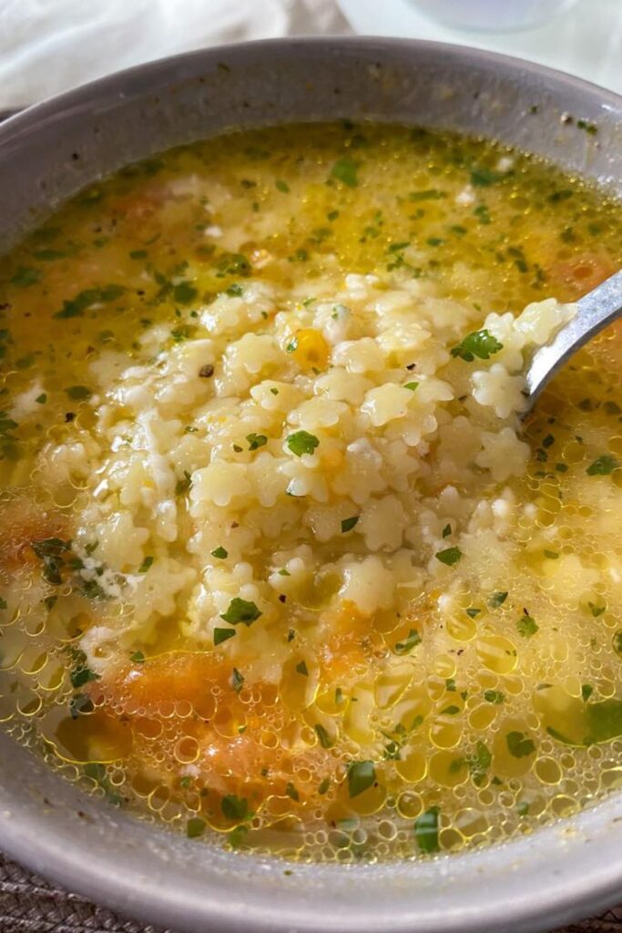 Close-up of Italian penicillin soup, featuring star-shaped pasta in a golden broth, garnished with fresh parsley. Comfort food in a grey bowl, ready to eat.