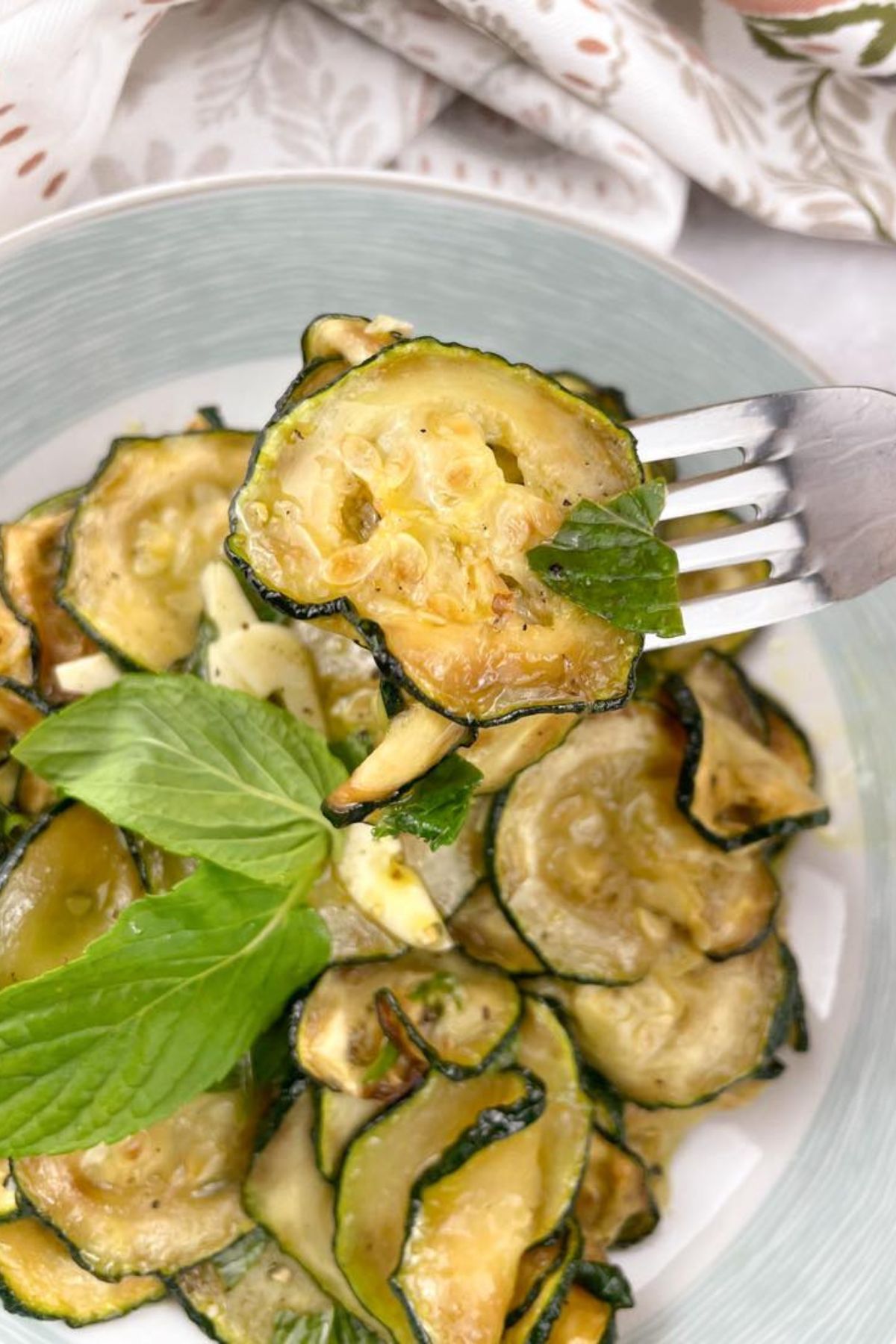 Zucchini alla scapece: A fork holds a glistening zucchini slice, showcasing the mint and vinegar dish. More zucchini, garnished with fresh mint, fills a light blue bowl.