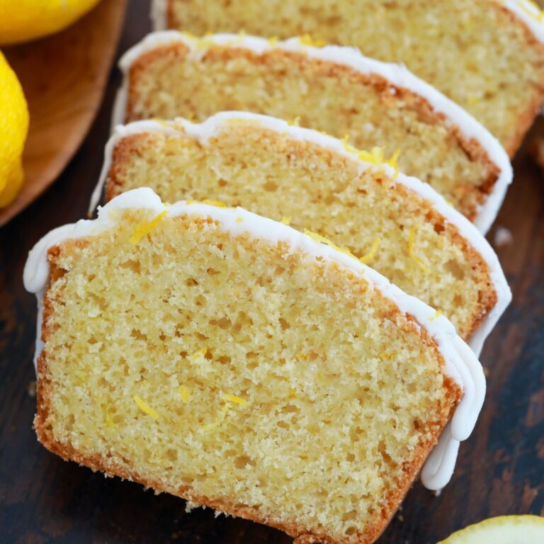 Slices of homemade lemon bread recipe with white icing and lemon zest garnish on a dark wood surface. Fresh lemons sit nearby.