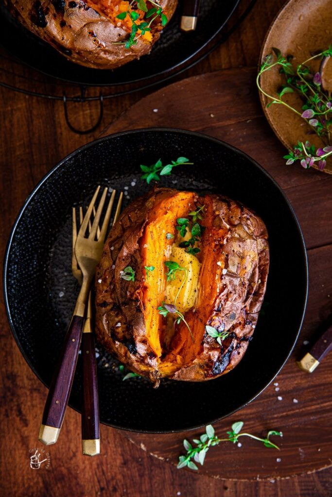 Baked sweet potato in black bowl with butter and herbs, rustic wooden background