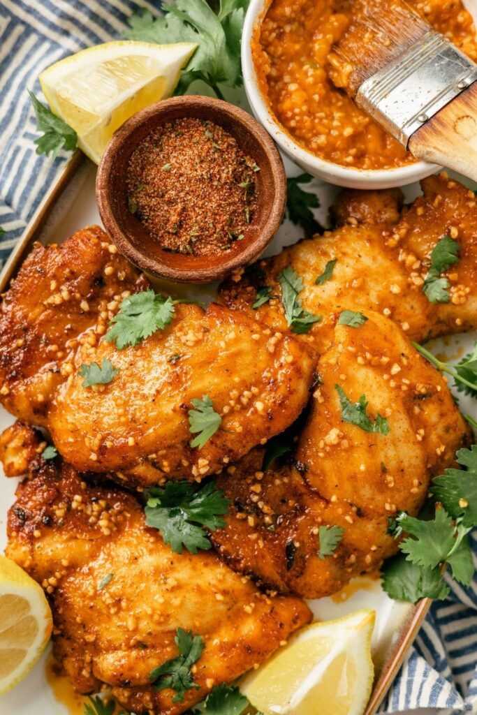 Glazed chicken thighs with lemon wedges, spice rub, and dipping sauce, garnished with cilantro.