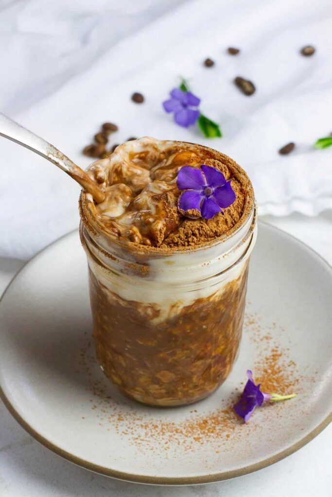 Tiramisu Overnight Oats Overnight oats recipe in a mason jar, topped with cream and a purple flower. Coffee beans surround the jar.
