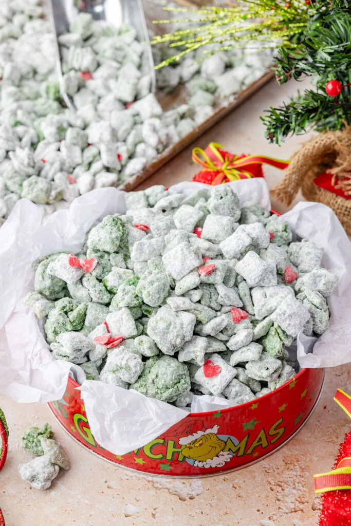Grinch Christmas Muddy Buddies in a red Grinch-themed tin. Festive green Chex mix puppy chow with powdered sugar and candy hearts, perfect for a Christmas potluck recipe.