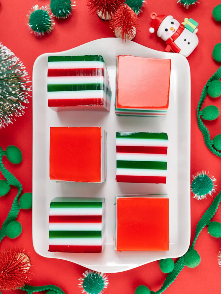 Festive Christmas potluck treats: gelatin squares in red, green, and white stripes and solids arranged on a white plate, surrounded by holiday decorations.