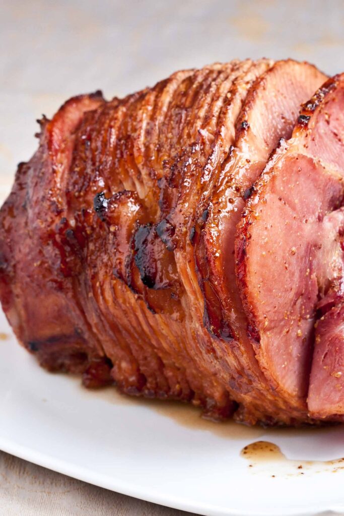 Glazed spiral-cut ham on a white plate, perfect for Christmas potluck recipes. The ham has a rich, caramelized glaze and is partially sliced, showing the tender meat inside.