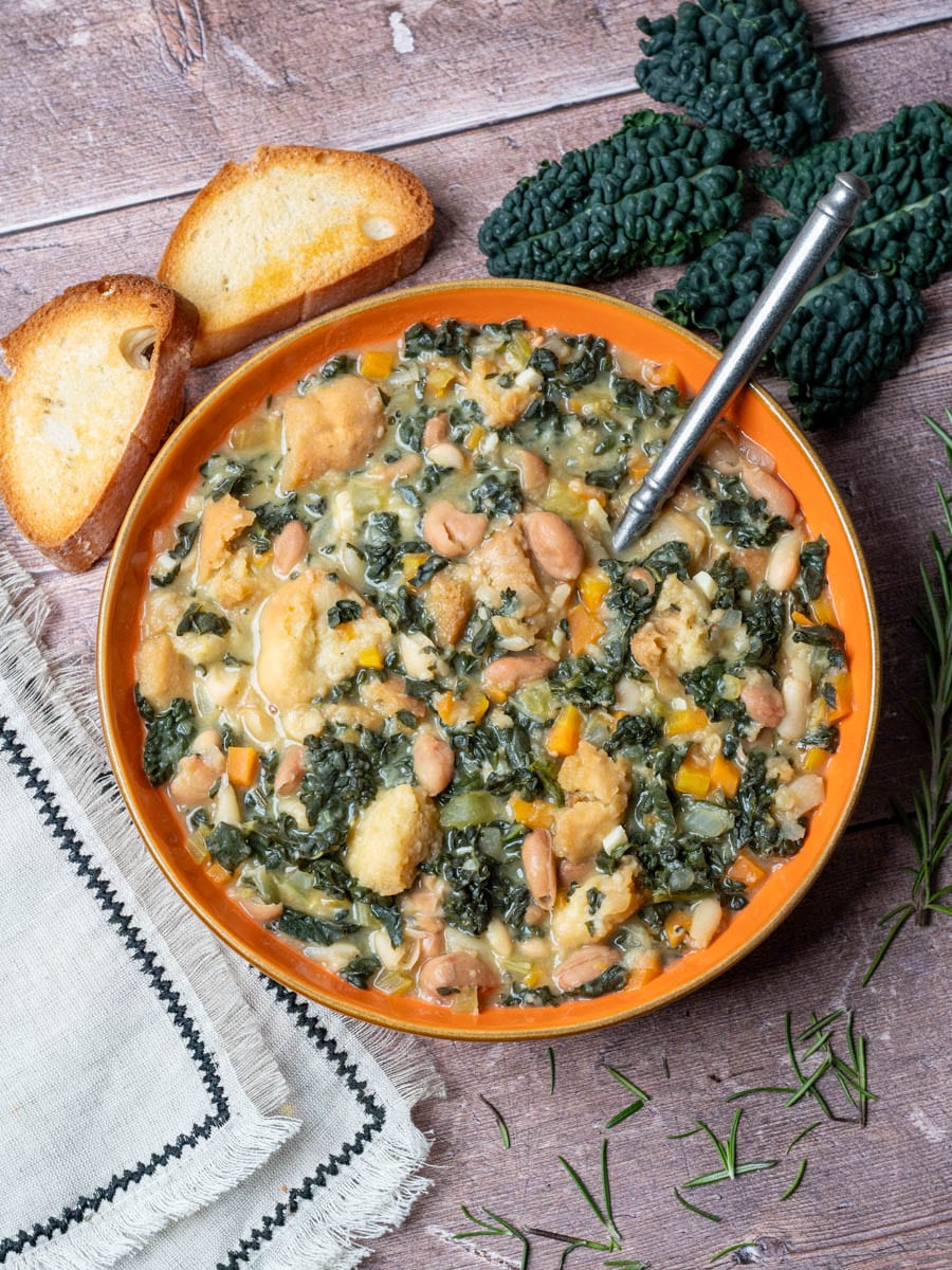 Hearty Tuscan ribollita soup, a delicious vegan Italian recipe, in an orange bowl with crusty bread. Featuring cavolo nero, cannellini beans, and rosemary, on a rustic wood table.