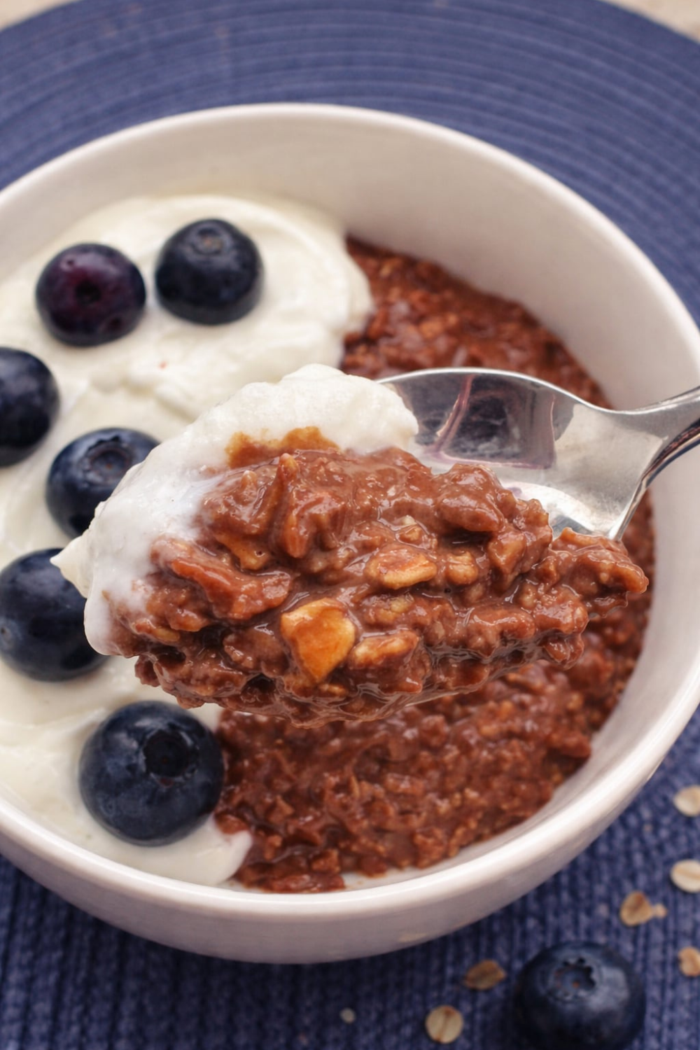 Spoonful of chocolate oatmeal with yogurt and blueberries, showing the texture of this quick, healthy breakfast. A bowl of chocolate oatmeal is in the background.