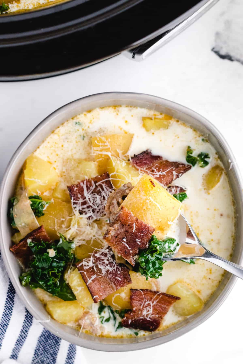 A bowl of creamy Italian slow cooker soup, topped with bacon, potatoes, kale, and parmesan cheese. A spoonful is lifted, showcasing the hearty texture of this Italian slow cooker recipe.