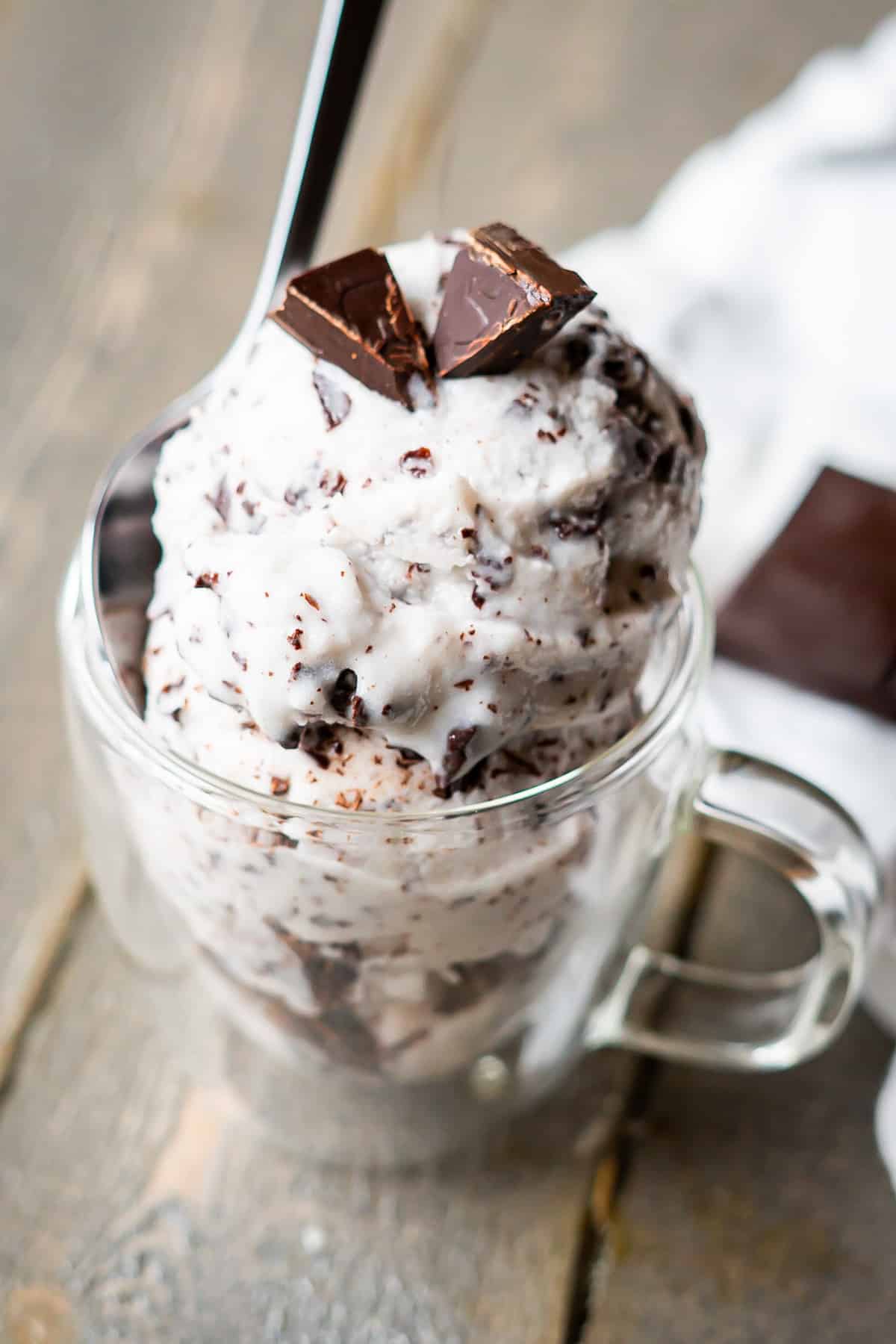 Vegan stracciatella ice cream in a glass mug, topped with dark chocolate pieces. A spoonful of creamy, chocolate chip dessert on a rustic wooden surface.