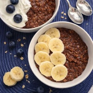 Two bowls of healthy chocolate oatmeal, one topped with yogurt and blueberries, the other with sliced bananas, on a blue mat with scattered oats.