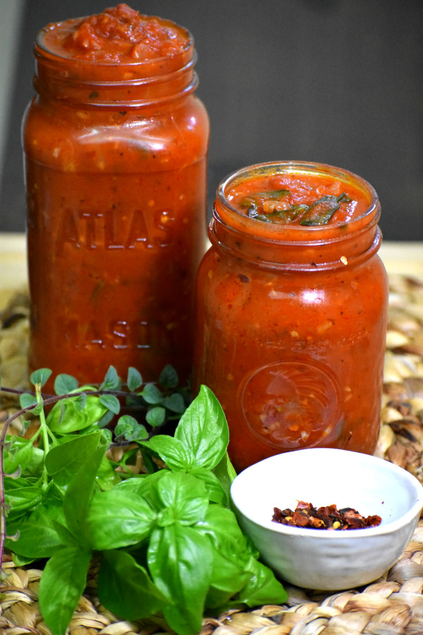 Two jars of homemade vegan Italian tomato sauce, garnished with fresh basil and red pepper flakes, sitting on a woven mat. Part of a collection of delicious vegan Italian recipes.