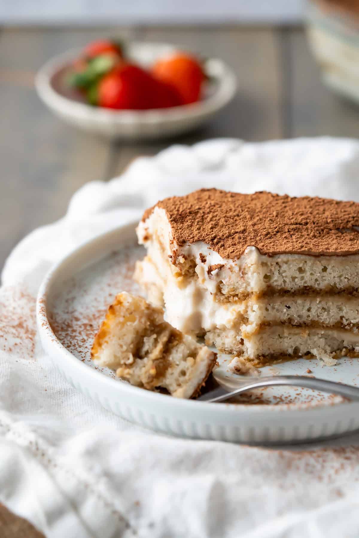 A slice of vegan tiramisu on a plate, dusted with cocoa powder, sits on a white cloth. Strawberries are blurred in the background. A delicious example of vegan Italian recipes.