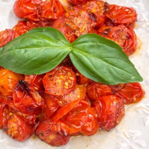 Air Fryer Roasted Tomatoes, glistening and caramelized, are piled on a white plate, topped with fresh basil leaves. The tomatoes are bright red and juicy, showcasing a quick and easy recipe.