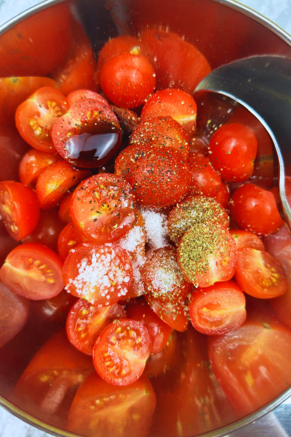 Fresh cherry tomatoes halved in a stainless steel bowl, seasoned with salt, pepper, herbs, and a drizzle of balsamic glaze.