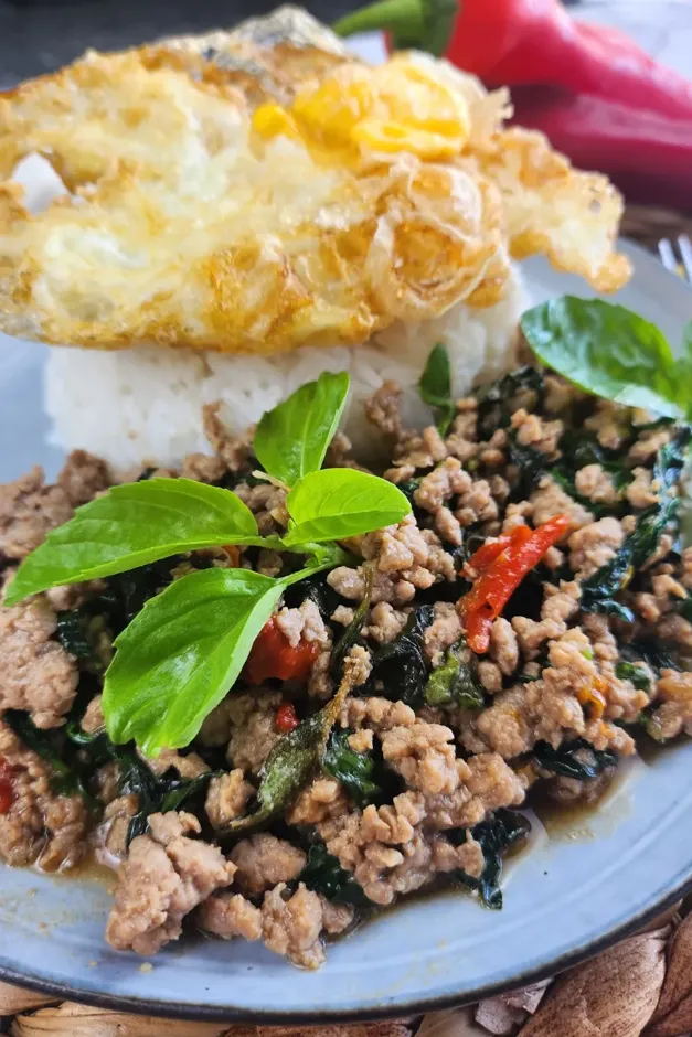 A close-up of a plate of Pad Krapow Moo, a popular Thai dish featuring stir-fried minced pork with holy basil, served over rice and topped with a crispy fried egg. This is one of many fresh basil recipes.