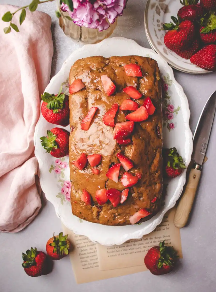 A loaf of glazed zucchini bread topped with fresh strawberry slices sits on a floral plate, surrounded by whole strawberries and a pink cloth.