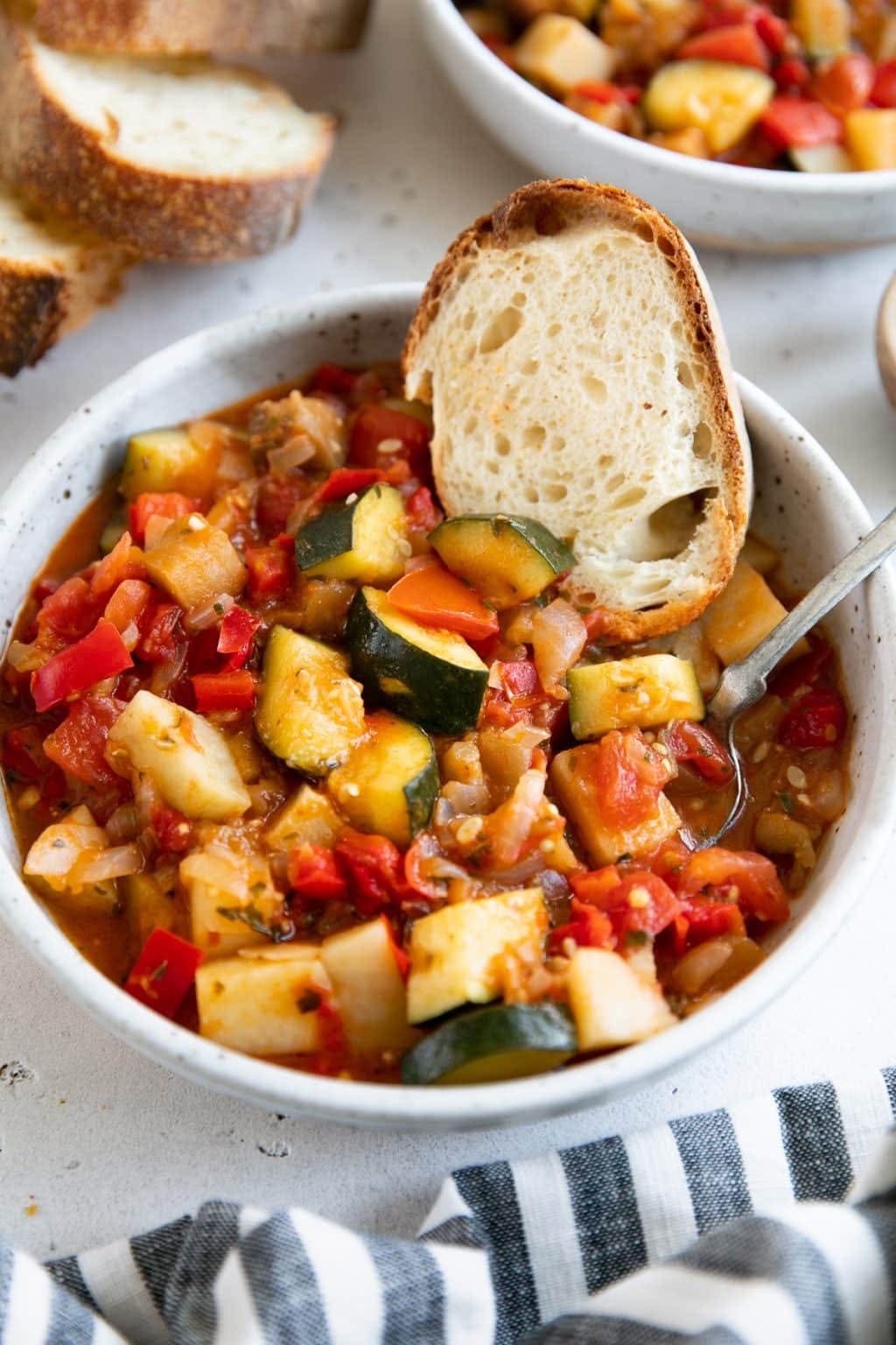 A rustic bowl of vibrant ratatouille, a delicious Italian gluten-free recipe, overflowing with zucchini, eggplant, tomatoes, and peppers, garnished with a slice of crusty bread.