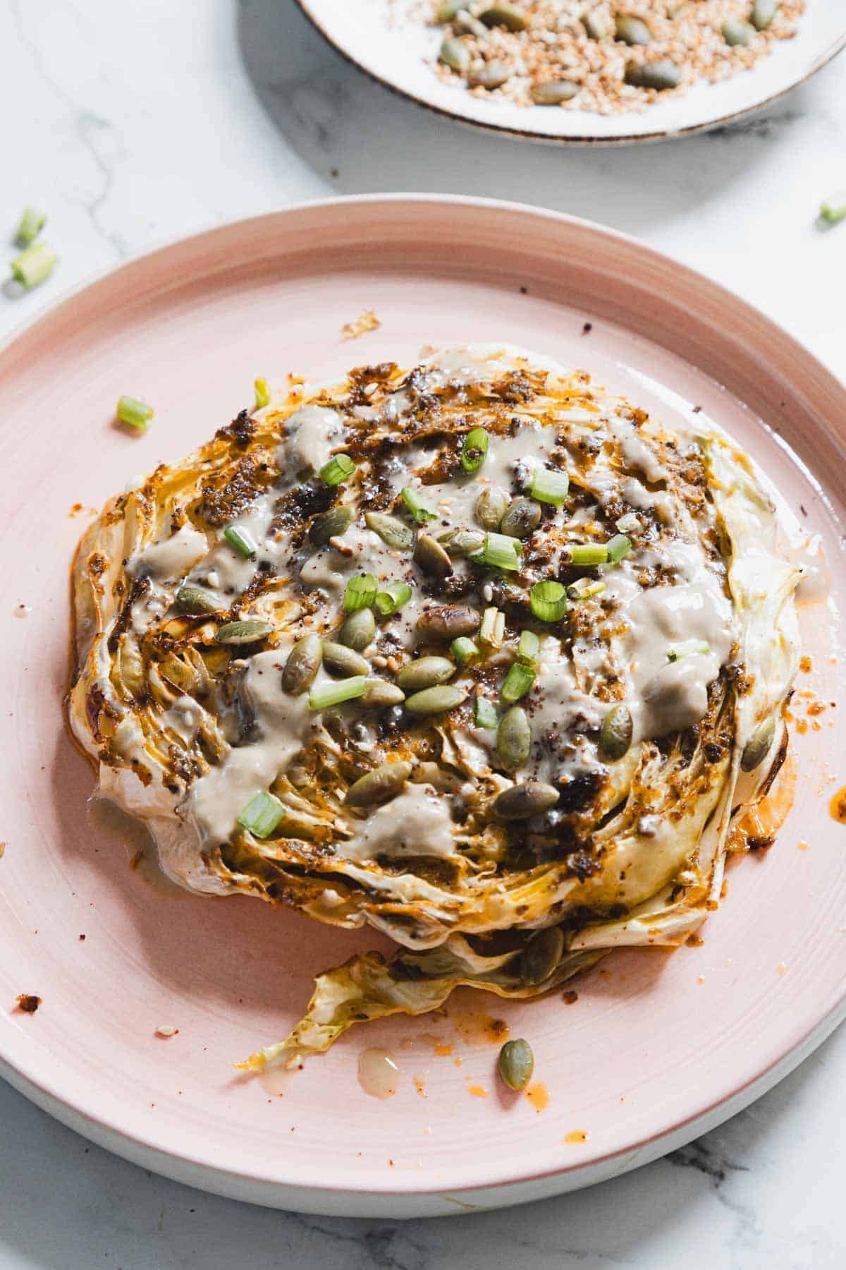 An air fryer cabbage steak topped with creamy tahini sauce, chopped green onions, and seeds, served on a pink plate. A small dish with seeds is visible in the background, all set on a sleek white marble table.