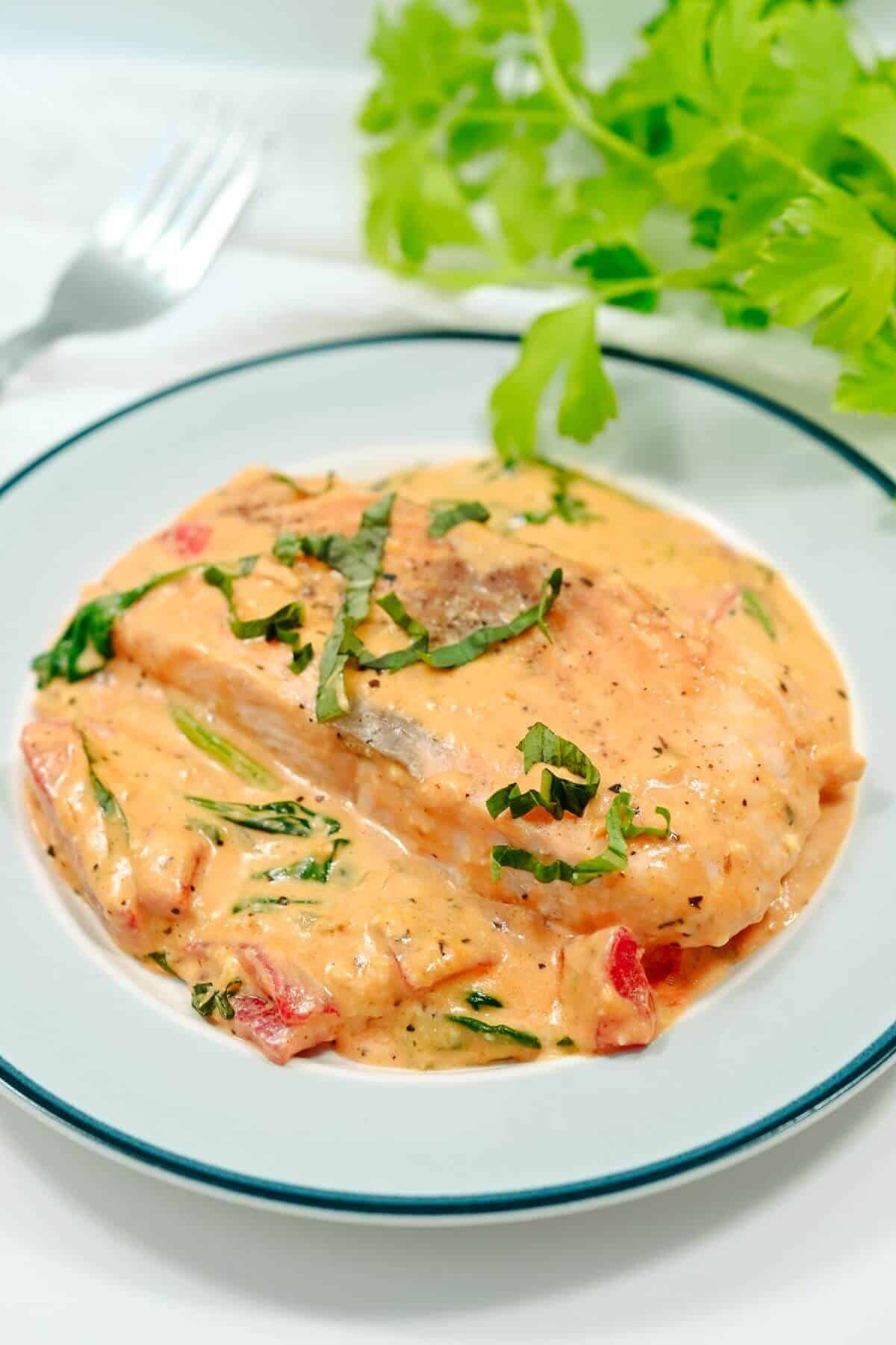 Plate of creamy sun-dried tomato chicken with herbs, spinach, and pepper slices, garnished with chopped basil, placed next to a sprig of fresh greens and a fork.